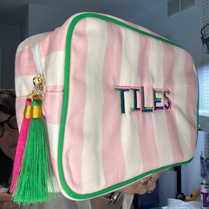 Pu&ograve; includere: Una borsa da viaggio rettangolare a righe rosa e bianche, con bordi verdi e cerniera dorata. La parola "TILES" &egrave; ricamata sul davanti in vari colori. Un nappa con dettagli rosa, verdi e dorati pende dalla cerniera.