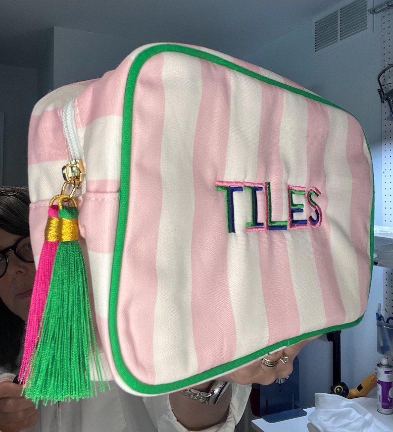 Pu&ograve; includere: Una borsa da viaggio rettangolare a righe rosa e bianche, con bordi verdi e cerniera dorata. La parola "TILES" &egrave; ricamata sul davanti in vari colori. Un nappa con dettagli rosa, verdi e dorati pende dalla cerniera.