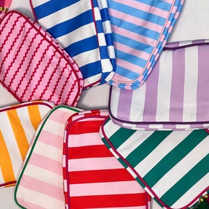 Pu&ograve; includere: Una collezione di trousse per cosmetici a righe colorate. Le borse presentano varie combinazioni di colori, tra cui rosa, blu, rosso, verde e arancione, con strisce bianche. Ogni borsa ha un bordo a contrasto e un nappa.
