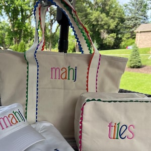 May include: A set of cream-colored tote bags, a cosmetic bag, and a baseball cap. The tote bag and cosmetic bag have rainbow-colored scalloped trim and embroidered text. The baseball cap has matching embroidery.