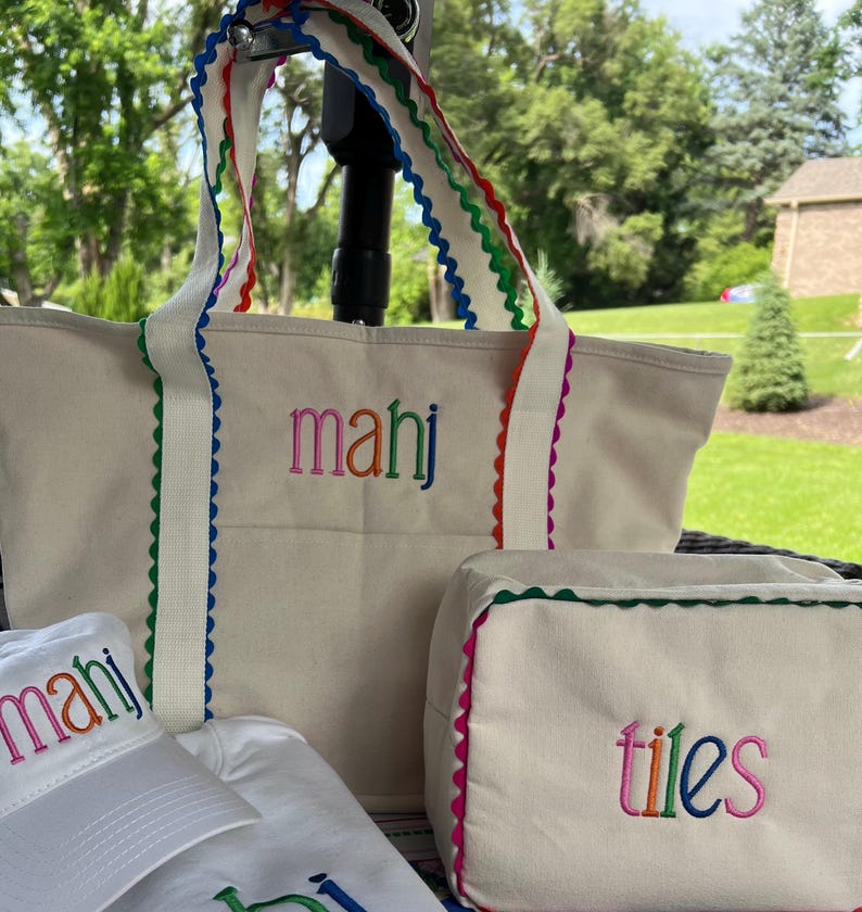 May include: A set of cream-colored tote bags, a cosmetic bag, and a baseball cap. The tote bag and cosmetic bag have rainbow-colored scalloped trim and embroidered text. The baseball cap has matching embroidery.
