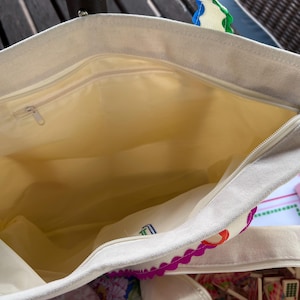 May include: Cream-colored tote bag with a zippered main compartment and a smaller interior pocket. The bag's handles have a scalloped edge design with red, orange, purple, and green accents. The interior lining is a light yellow color.