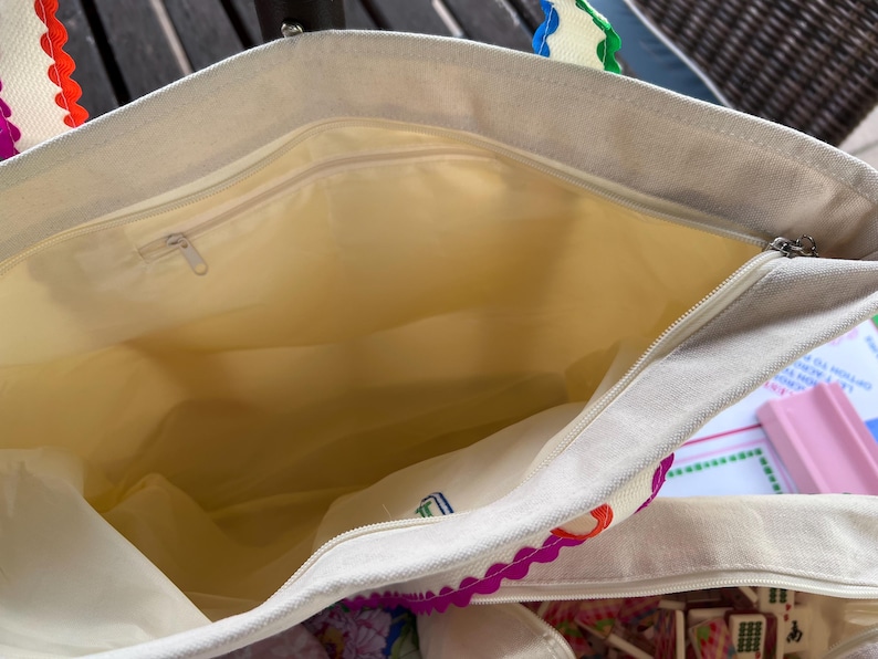 May include: Cream-colored tote bag with a zippered main compartment and a smaller interior pocket. The bag's handles have a scalloped edge design with red, orange, purple, and green accents. The interior lining is a light yellow color.