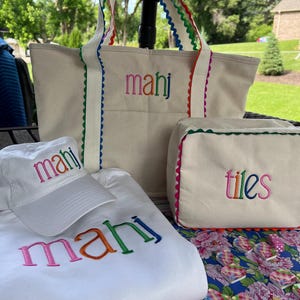 May include: A collection of white canvas tote bags, a hat, and a sweatshirt, all embroidered with the name "mahj" in rainbow colors. The tote bags and cosmetic bag have colorful scalloped trim. The cosmetic bag is embroidered with the word "tiles".