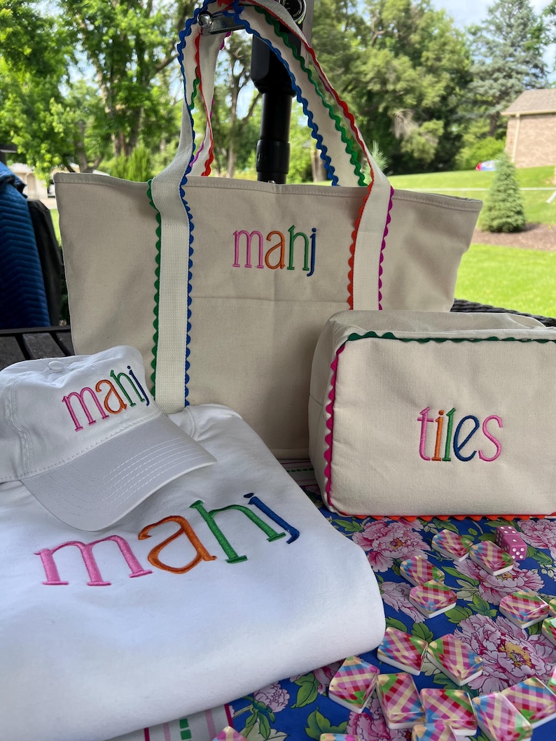 May include: A collection of white canvas tote bags, a hat, and a sweatshirt, all embroidered with the name "mahj" in rainbow colors. The tote bags and cosmetic bag have colorful scalloped trim. The cosmetic bag is embroidered with the word "tiles".