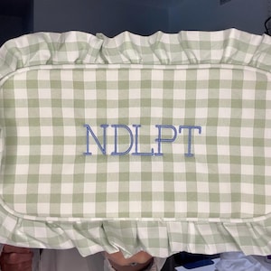 Può includere: Un cuscino rettangolare con motivo a quadretti verde e bianco. Il cuscino ha un bordo a balze e lettere blu ricamate che leggono "NDLPT". Un oggetto decorativo.