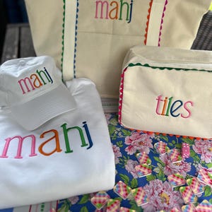 May include: A collection of cream-colored tote bags, a baseball cap, and a sweatshirt, all embroidered with the word "mahj" in colorful letters. A matching cream-colored cosmetic bag with the word "tiles" is also present. The items are arranged on a floral patterned surface.