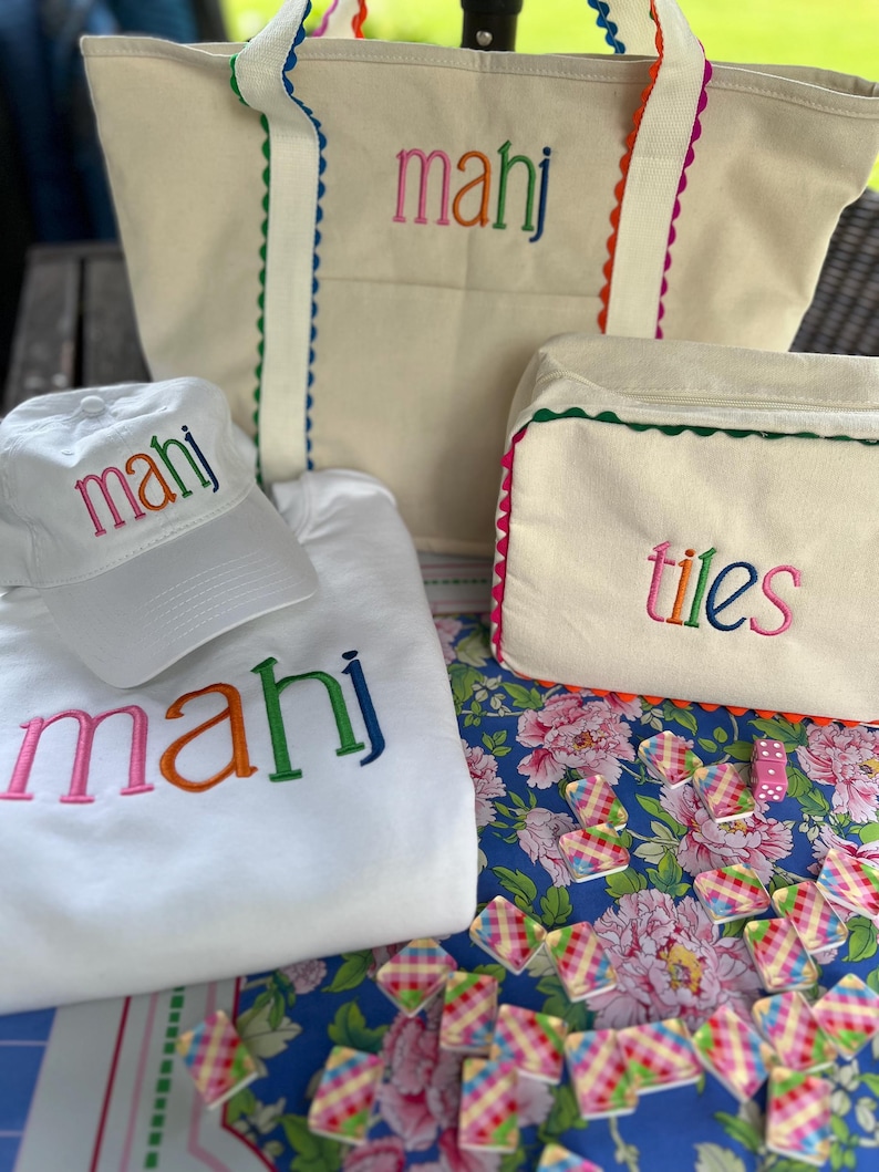 May include: A collection of cream-colored tote bags, a baseball cap, and a sweatshirt, all embroidered with the word "mahj" in colorful letters. A matching cream-colored cosmetic bag with the word "tiles" is also present. The items are arranged on a floral patterned surface.
