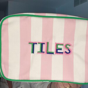 Pu&ograve; includere: Pochette per cosmetici a righe rosa e bianche con finiture verdi e nappa verde. La parola "TILES" &egrave; ricamata a lettere colorate sul davanti. La borsa &egrave; rettangolare e sembra essere in tessuto.