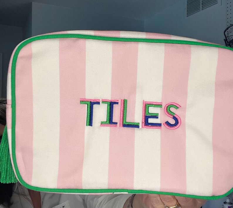 Pu&ograve; includere: Pochette per cosmetici a righe rosa e bianche con finiture verdi e nappa verde. La parola "TILES" &egrave; ricamata a lettere colorate sul davanti. La borsa &egrave; rettangolare e sembra essere in tessuto.