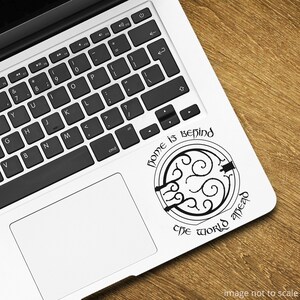 May include: A laptop computer with a black and white sticker on the back of the trackpad. The sticker features a circular design with a quote that reads "Home is behind, the world ahead."