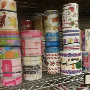 May include: Assortment of colorful rolls of decorative paper with various patterns and designs. The rolls are stacked on a metal shelf. Some of the designs include leaves, polka dots, stripes, and holiday themes.
