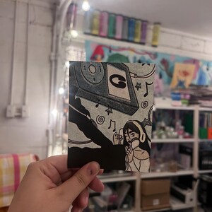 May include: A rectangular art print featuring a black and white illustration of a person wearing headphones and singing into a microphone. Above the person, a cassette tape is depicted with a letter G on the label, surrounded by musical notes.