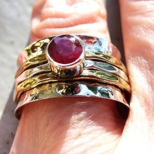 以下が含まれることがあります： 赤いルビーの宝石が付いた銀と金のリング。リングは、複数のバンドで重ねられるデザインです。
