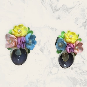 Op de afbeelding: Een paar schelpoorbellen. Elke oorbel heeft een cluster van kleurrijke bloemen in geel, roze, paars en blauw, met een parel in het midden. De bloemen zijn bevestigd aan een donkerpaarse schelpbasis.