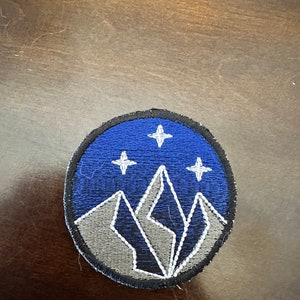 May include: A blue and gray embroidered patch with a mountain range and three white stars. The patch is circular and has a black outline.