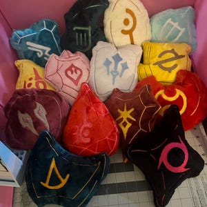 May include: A collection of plush pillows in various shapes and colors, each embroidered with unique symbols. The pillows are in shades of blue, pink, yellow, red, and black, and are arranged inside a pink storage bin. The symbols are in gold, white, and pink.