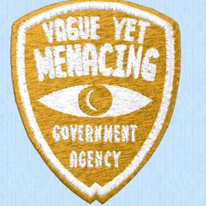 May include: A gold embroidered patch with a white outline in the shape of a shield. The patch features a white eye with a crescent moon inside, and the text "VAGUE YET MENACING GOVERNMENT AGENCY".