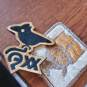 May include: Two embroidered patches. One is black with a gold outline and depicts a stylized bird with the word "Pax" in gold. The other is beige with a gold outline and depicts a stylized owl with the word "Moon" in gold.