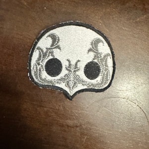 May include: A white embroidered patch with a black skull design. The skull has two black eyes and a detailed design around the eyes and mouth.
