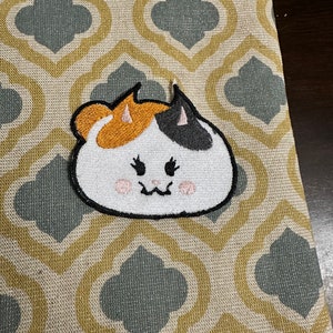 May include: A white, orange, and black embroidered patch of a cartoon cat with pink cheeks. The cat is smiling and has a black nose.