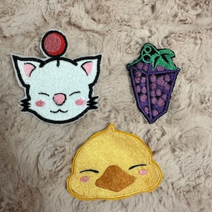 May include: Three embroidered patches featuring cartoon animal faces. A white cat with a red ball on its head, a yellow chick, and a purple grape.
