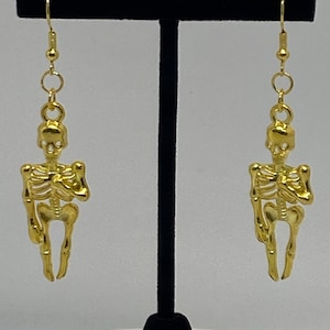 May include: A pair of gold skeleton earrings. The earrings are hanging from a black display stand.