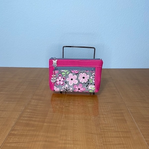 May include: A pink zippered pouch with a floral pattern in pink, gray, and green. The pouch has a gray zipper pocket and a silver charm. The pouch is on a black metal stand.