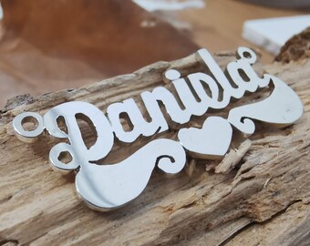 Könnte beinhalten: Silberne Namensschilderkette mit dem Namen "Daniela" in einer kursivschriftlichen Schriftart mit zwei Herzen. Die Kette liegt auf einer Holzoberfläche.