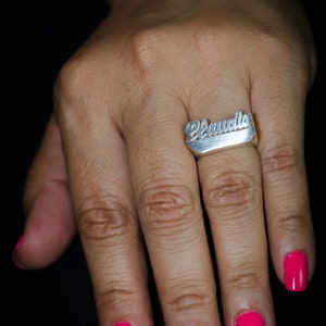 K&ouml;nnte beinhalten: Ein silberner Ring mit dem Namen "Claudia" in Schreibschrift. Der Ring befindet sich an einem Finger mit leuchtend pinkfarbenem Nagellack. Der Hintergrund ist schwarz.