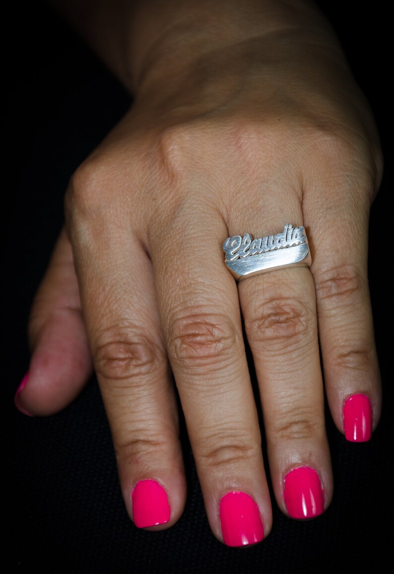K&ouml;nnte beinhalten: Ein silberner Ring mit dem Namen "Claudia" in Schreibschrift. Der Ring befindet sich an einem Finger mit leuchtend pinkfarbenem Nagellack. Der Hintergrund ist schwarz.