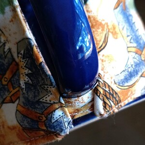 Puede incluir: Una taza de cer&aacute;mica azul con asa, envuelta en una funda de tela con un patr&oacute;n de botas de vaquero. La tela es blanca con detalles marrones y azules.