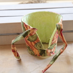 Puede incluir: Un calientavasos de tela verde y marr&oacute;n con un forro verde. El calientavasos tiene dos lazos que se pueden atar alrededor de una taza.