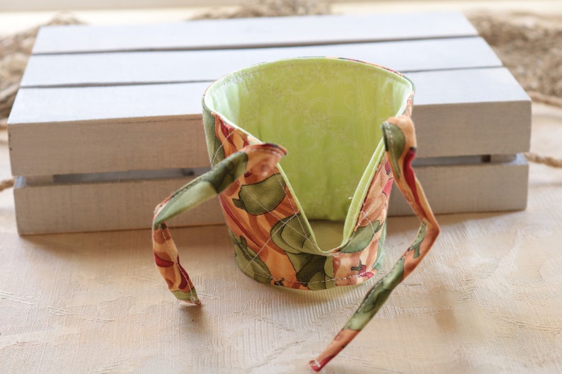 Puede incluir: Un calientavasos de tela verde y marr&oacute;n con un forro verde. El calientavasos tiene dos lazos que se pueden atar alrededor de una taza.