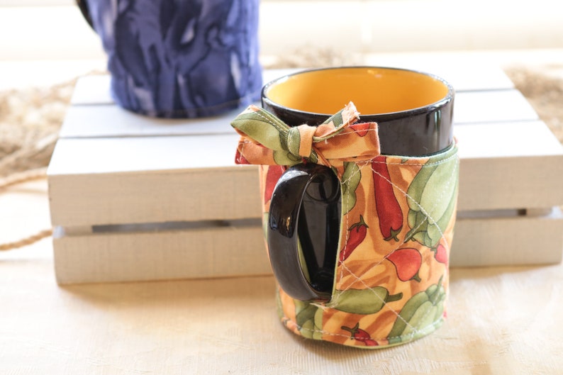 Puede incluir: Una taza negra con un borde amarillo y una funda de tela con un patr&oacute;n de pimientos rojos y verdes sobre un fondo beige. La funda tiene un lazo verde y beige atado alrededor del asa.