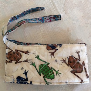 Puede incluir: Una bolsa de tela acolchada blanca con un dise&ntilde;o de rana. La bolsa tiene una correa de tela azul y verde con un patr&oacute;n de cachemira. La bolsa mide aproximadamente 20 cm de largo y 8 cm de ancho.