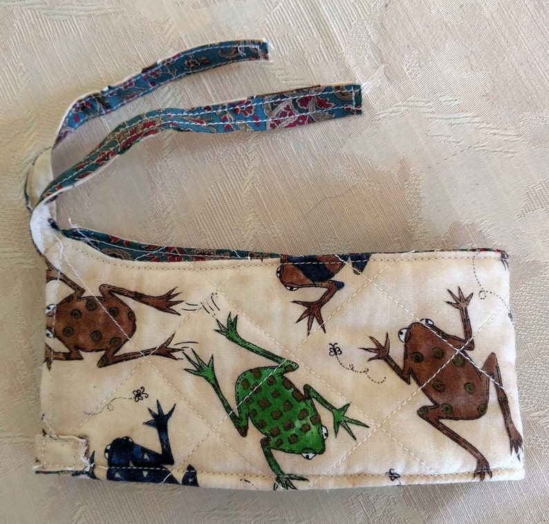 Puede incluir: Una bolsa de tela acolchada blanca con un dise&ntilde;o de rana. La bolsa tiene una correa de tela azul y verde con un patr&oacute;n de cachemira. La bolsa mide aproximadamente 20 cm de largo y 8 cm de ancho.