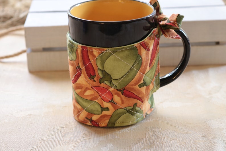 Puede incluir: Una taza negra con interior amarillo est&aacute; envuelta en un calientavasos de tela. El calientavasos tiene un patr&oacute;n de pimientos verdes y chiles rojos sobre un fondo marr&oacute;n.