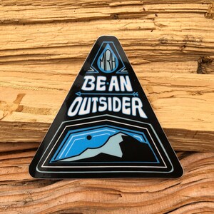 May include: A black triangular sticker with a blue mountain scene and the text "BE AN OUTSIDER" in white. The sticker also has a blue drop with the letters "MRH" inside.