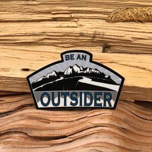 May include: A black and white sticker with a mountain range design and the text "BE AN OUTSIDER" in blue lettering.