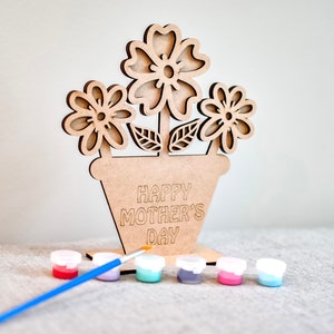 Puede incluir: Kit de manualidades de madera con una maceta con tres flores y el texto "Happy Mother's Day". El kit incluye un pincel y cinco pequeños botes de pintura.