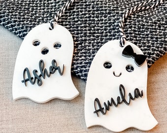 Op de afbeelding: Twee witte spookvormige labels met zwarte accenten en namen. Op één label staat "Asher" en op de andere "Amelia". Het Amelia-label heeft een zwarte strik. Beide labels hebben een zwart-wit touwtje.