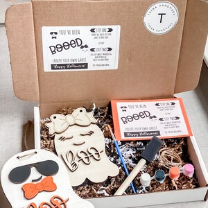 May include: A craft kit for creating your own ghost decorations. The kit includes two unfinished wooden ghost shapes, paint, brushes, and instructions. The instructions say "You've Been Booed!" and "Create Your Own Ghost! Happy Halloween!"
