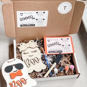May include: A white box with a brown lid is open, revealing a craft kit for creating your own ghost. The kit includes two wooden ghost shapes, paint, brushes, and instructions. The box is labeled "You've Been Booed!" and "Create Your Own Ghost".