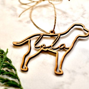 May include: A wooden dog ornament with the name "Lola" cut out inside the silhouette. The ornament is hanging from a twine string.
