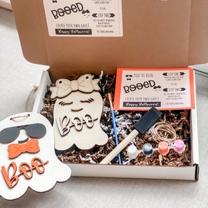 May include: A craft kit for making a ghost decoration. The kit includes two unfinished wooden ghost shapes, paint, a paintbrush, twine, and instructions. The instructions say "You've been Booed! Create your own ghost! Happy Halloween!"