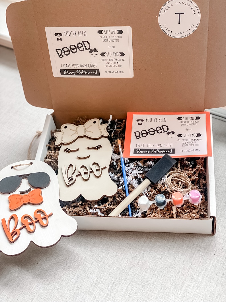 May include: A craft kit for making a ghost decoration. The kit includes two unfinished wooden ghost shapes, paint, a paintbrush, twine, and instructions. The instructions say "You've been Booed! Create your own ghost! Happy Halloween!"