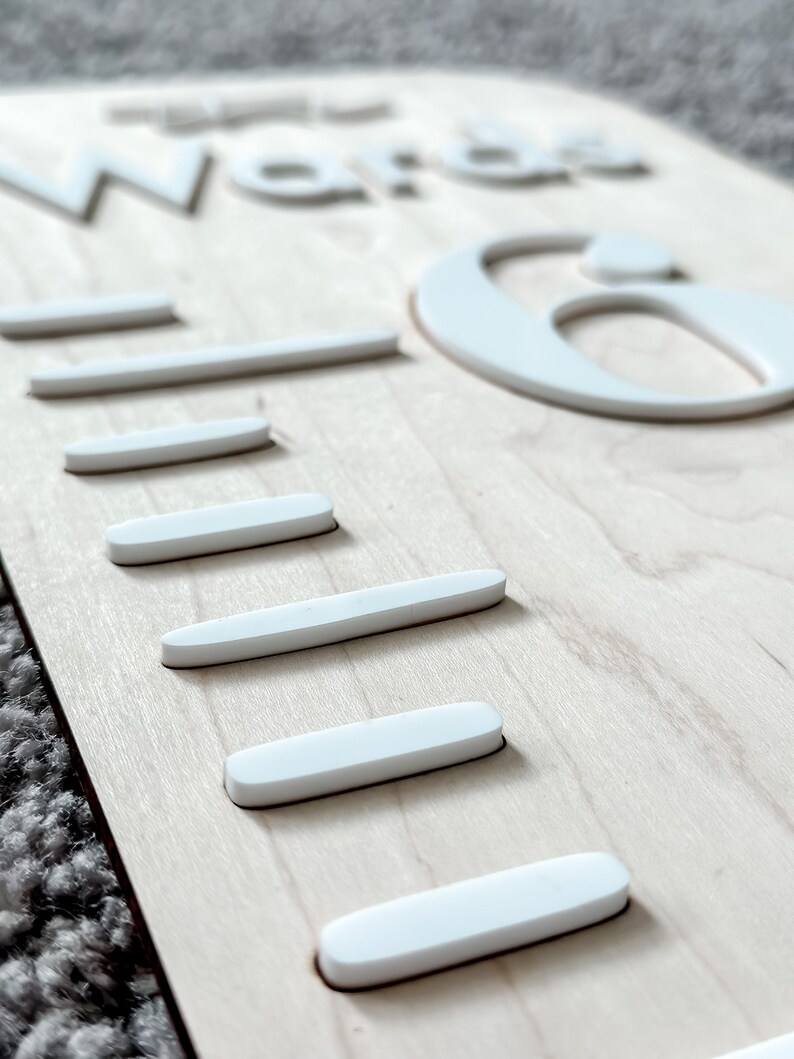 May include: A wooden growth chart with the words "The Word is" and the number "6" in white raised letters. The chart has five white lines for measuring height.