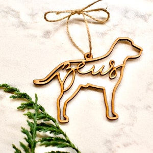 May include: Wooden dog-shaped Christmas ornament with the name "Zeus" cut out inside the silhouette. The ornament is hanging from a twine string with a bow.