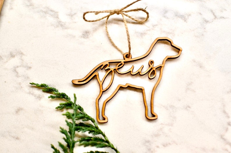May include: Wooden dog-shaped Christmas ornament with the name "Zeus" cut out inside the silhouette. The ornament is hanging from a twine string with a bow.